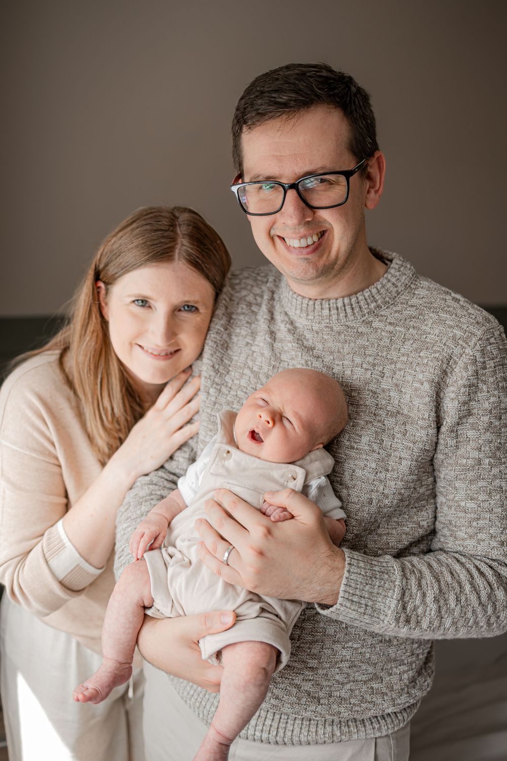 Smiling parents holding their newborn baby, posing together for a family portrait in a warm, neutral-toned setting.