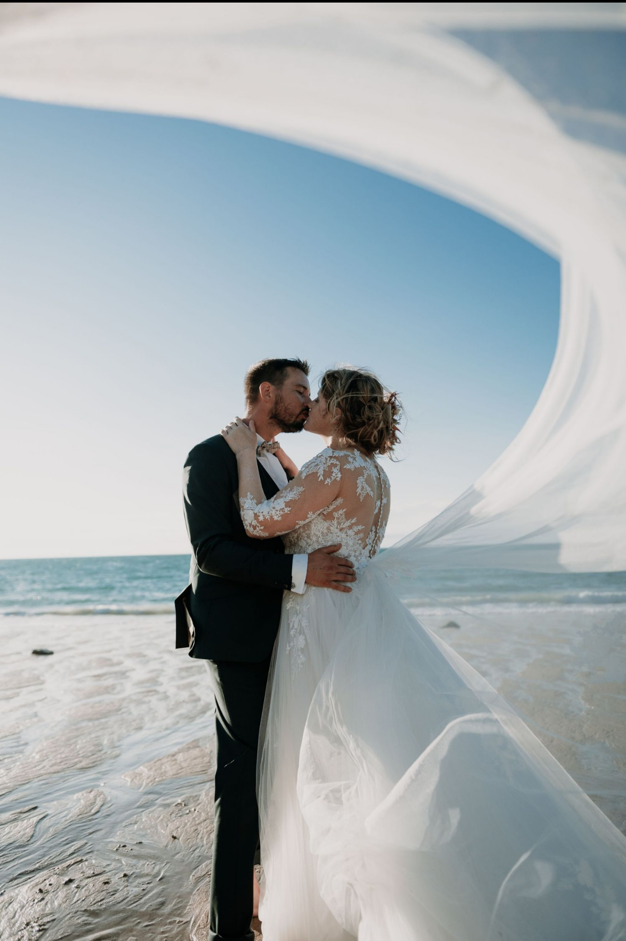 Couple de mariés s’embrassant au coucher du soleil sur une plage en Bretagne, séance photo de mariage bord de mer avec voile au vent