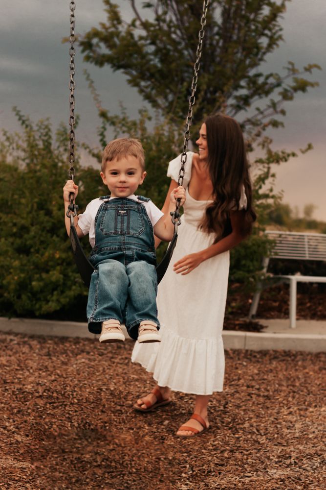 A smiling mother in a white dress pushes her toddler boy on a playground swing outdoors. | Westminster Colorado Family Photographer