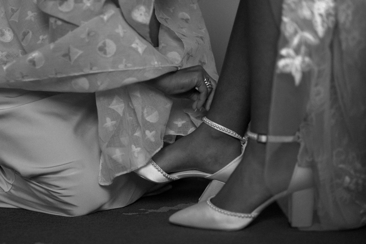A black and white detail shot of an elegant ankle strap heel being fastened during a formal occasion.