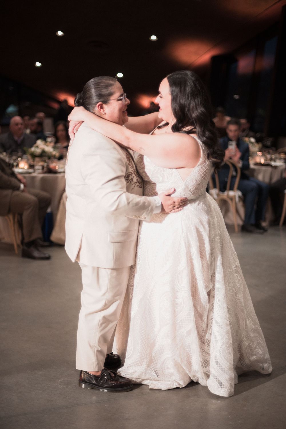 Bride in lace off-shoulder gown shares first dance with partner in cream suit at elegant wedding reception.