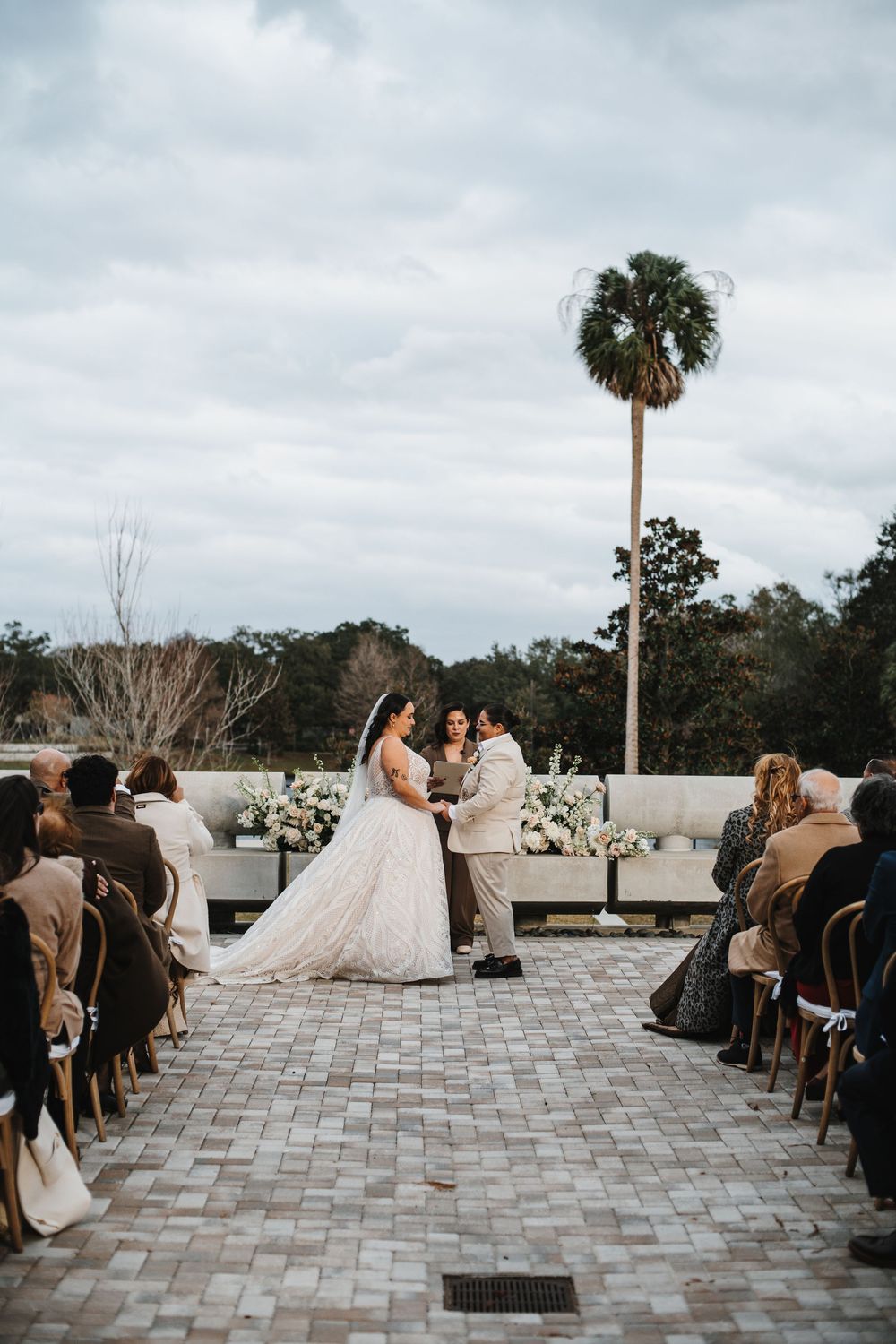 Wedding ceremony guests seated on a stone patio overlooking palm trees and cloudy skies during an outdoor celebration.