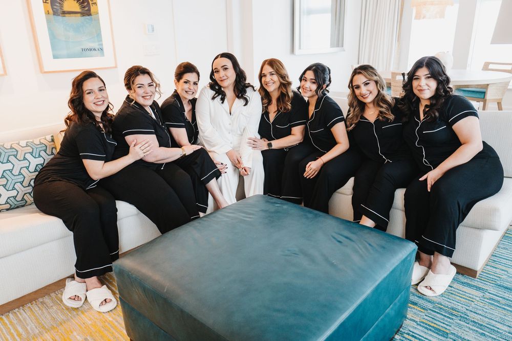 A group in matching black and white outfits sits together on a white couch around a teal ottoman in a bright room.