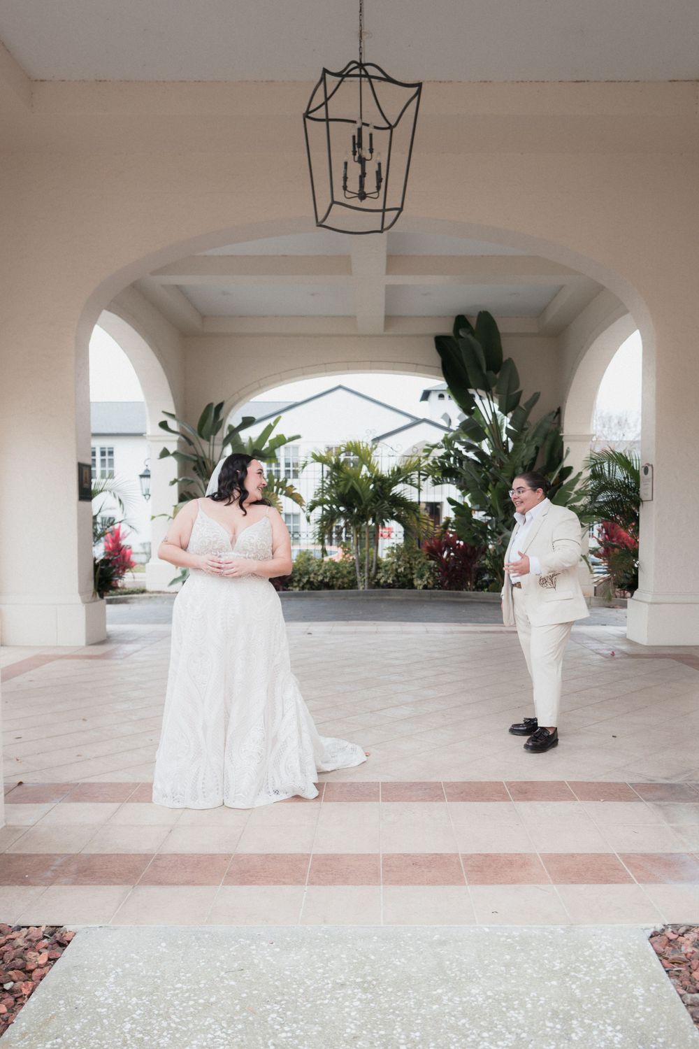 Bride in white gown and groom in cream suit share a first look moment under elegant arched colonnade with tropical greenery.