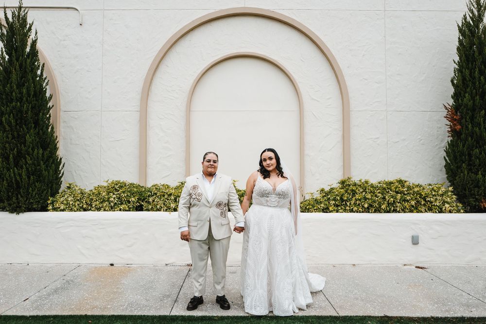 Wedding couple poses in front of white arched wall with cypress trees for elegant outdoor portraits.