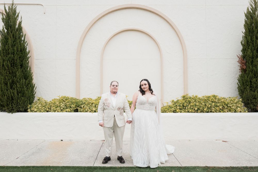 Two brides in white wedding attire hold hands in front of an elegant arched wall with cypress trees.
