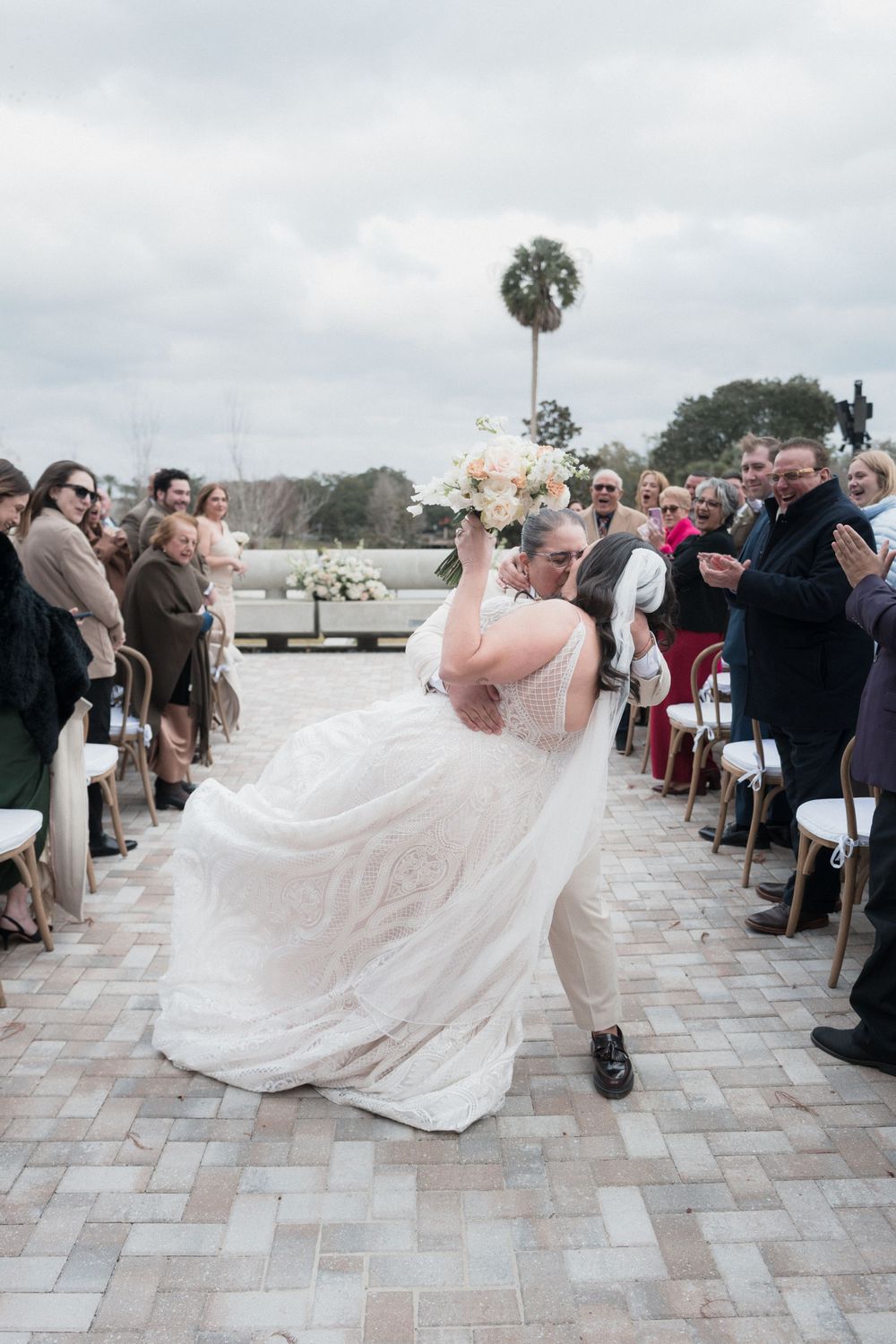 Bride and groom share a dip kiss while walking down the aisle at an outdoor wedding ceremony with guests applauding.
