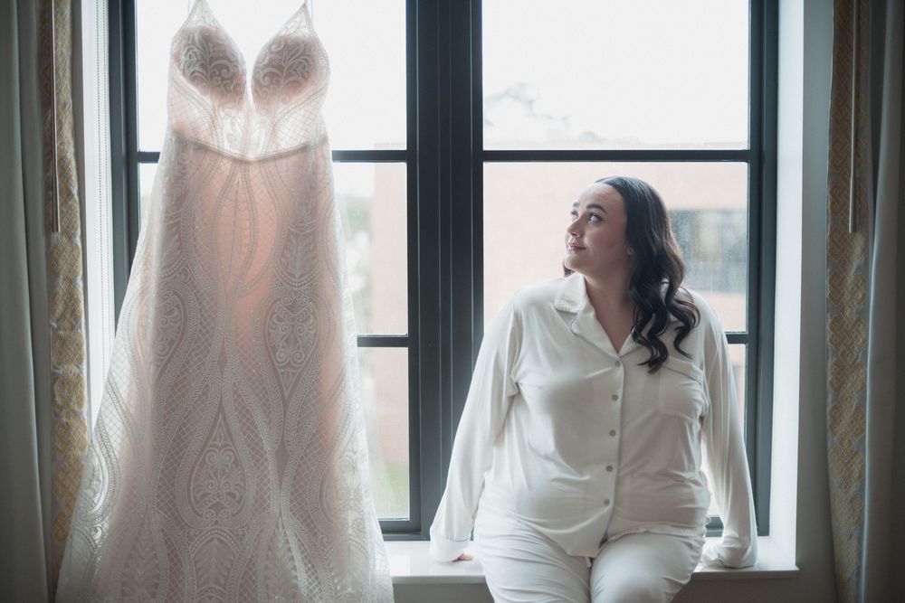 Bride in white pajamas sitting by window admiring her lace wedding dress hanging in natural light.