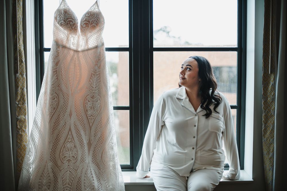 A detailed lace wedding dress hangs by a window while someone in white loungewear sits nearby looking peaceful.