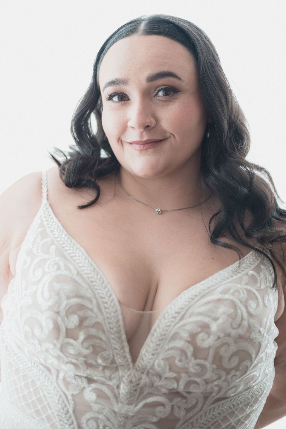 Woman in white lace deep V-neck bridal gown with delicate necklace, smiling against light background.