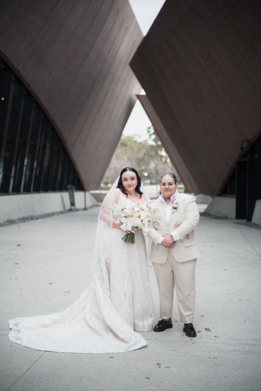 Two brides pose in wedding attire before geometric wooden architecture, one in white gown with bouquet, one in cream suit.