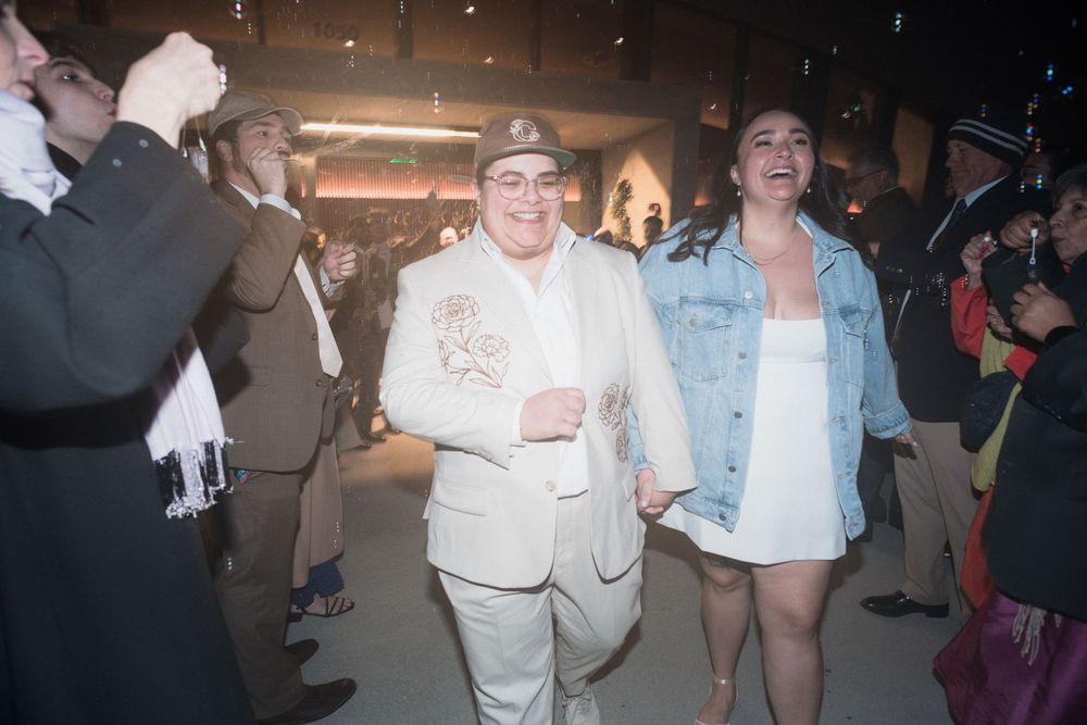 Two people walk hand-in-hand through a celebratory crowd at an indoor event, one wearing a white embroidered blazer.