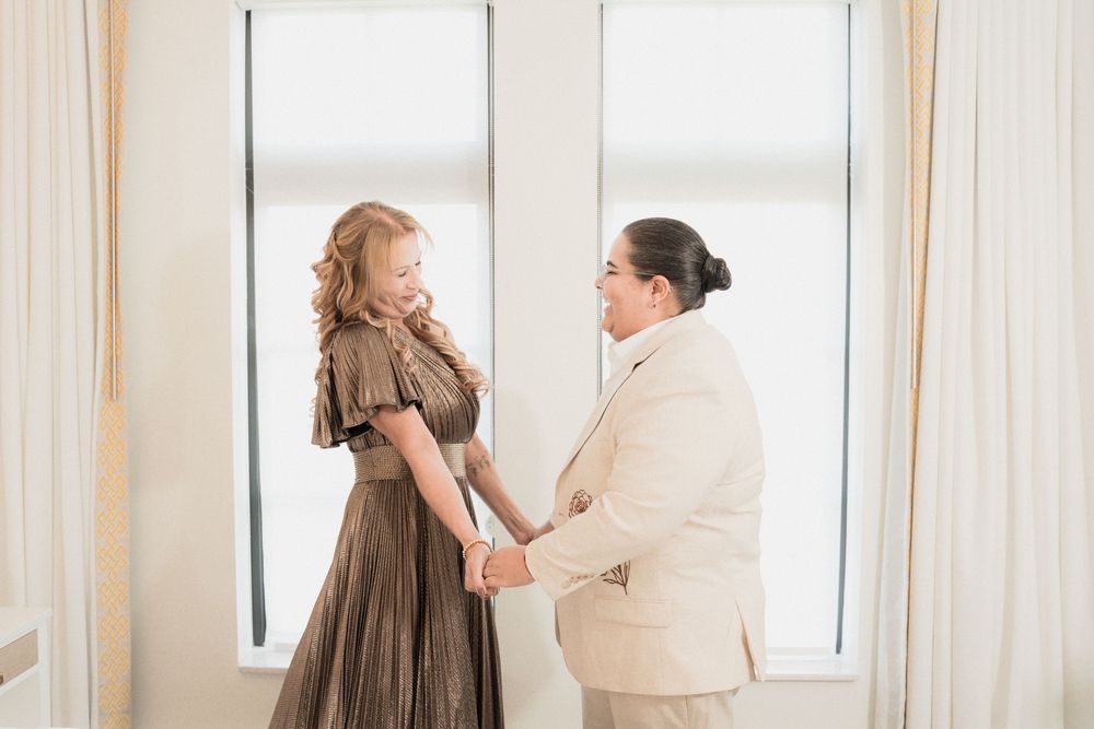 Couple holding hands and smiling at each other in a bright white room, woman in brown dress, man in beige suit.