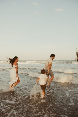 Family of three playing in ocean waves at sunset near a beach pier.