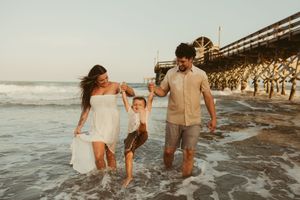 Happy family swinging young child between them while walking on a sunny beach near a pier.