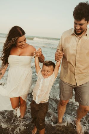 Smiling parents holding their young son's hands as waves splash around them on the beach.