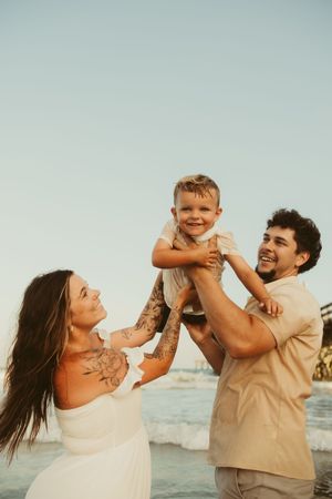 Laughing father lifting toddler son high in the air while mother watches on a sunny beach.