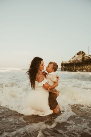 Mother kneeling in ocean waves holding her young son while smiling at each other near a pier.