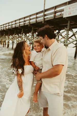 Happy family with young boy laughing together in ocean waves near a wooden pier.