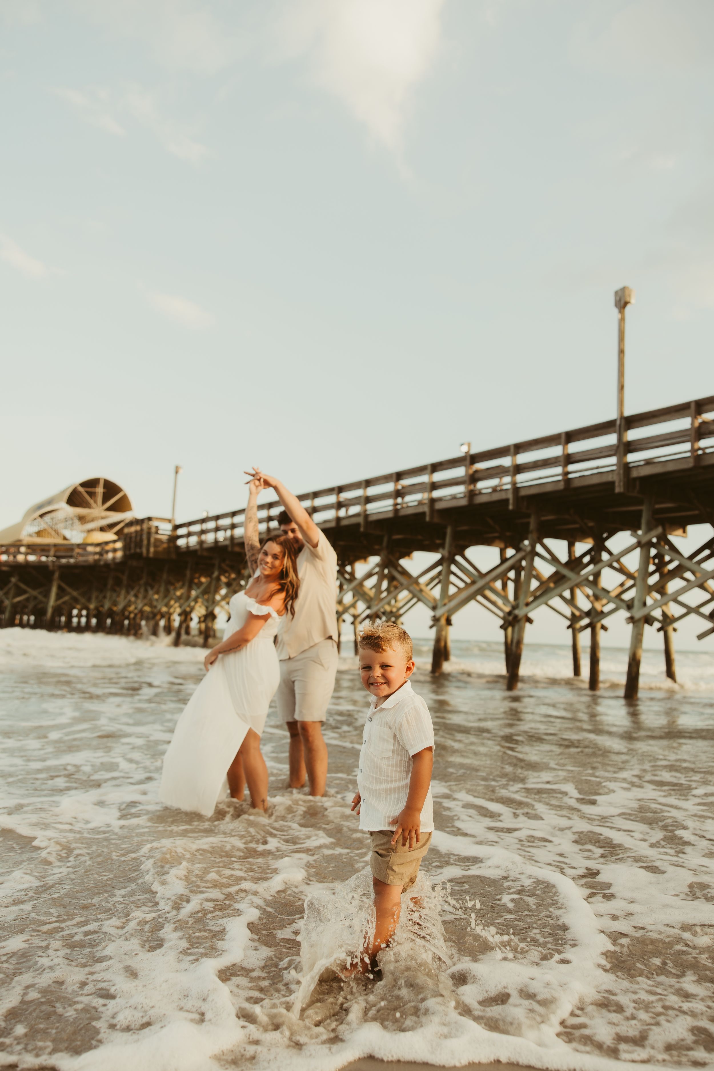Joyful boy splashing in ocean waves as parents dance behind him near a beach pier.