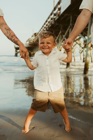 Laughing toddler boy holding parents hands on sandy beach near a wooden pier at sunset.