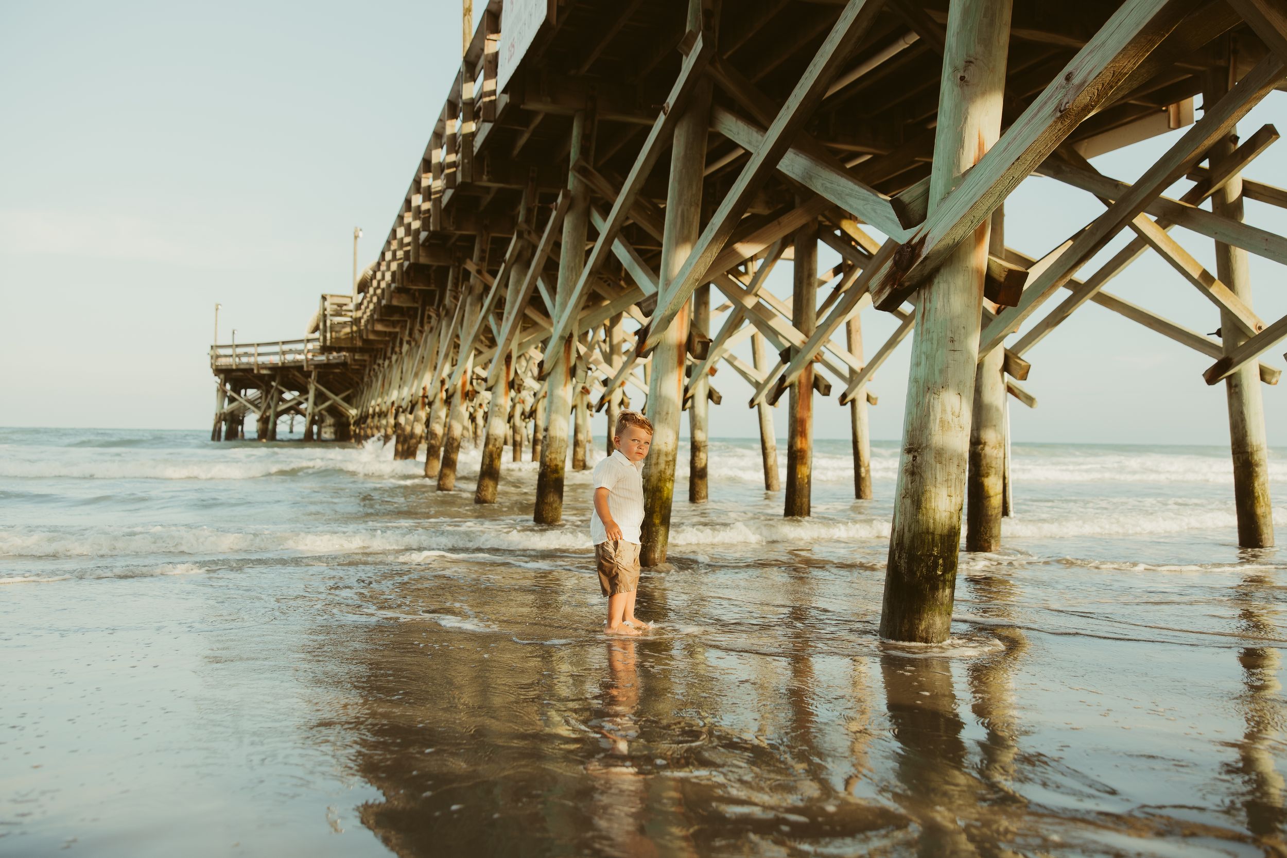 Woman standing under a wooden beach pier with waves washing over her feet at golden hour.