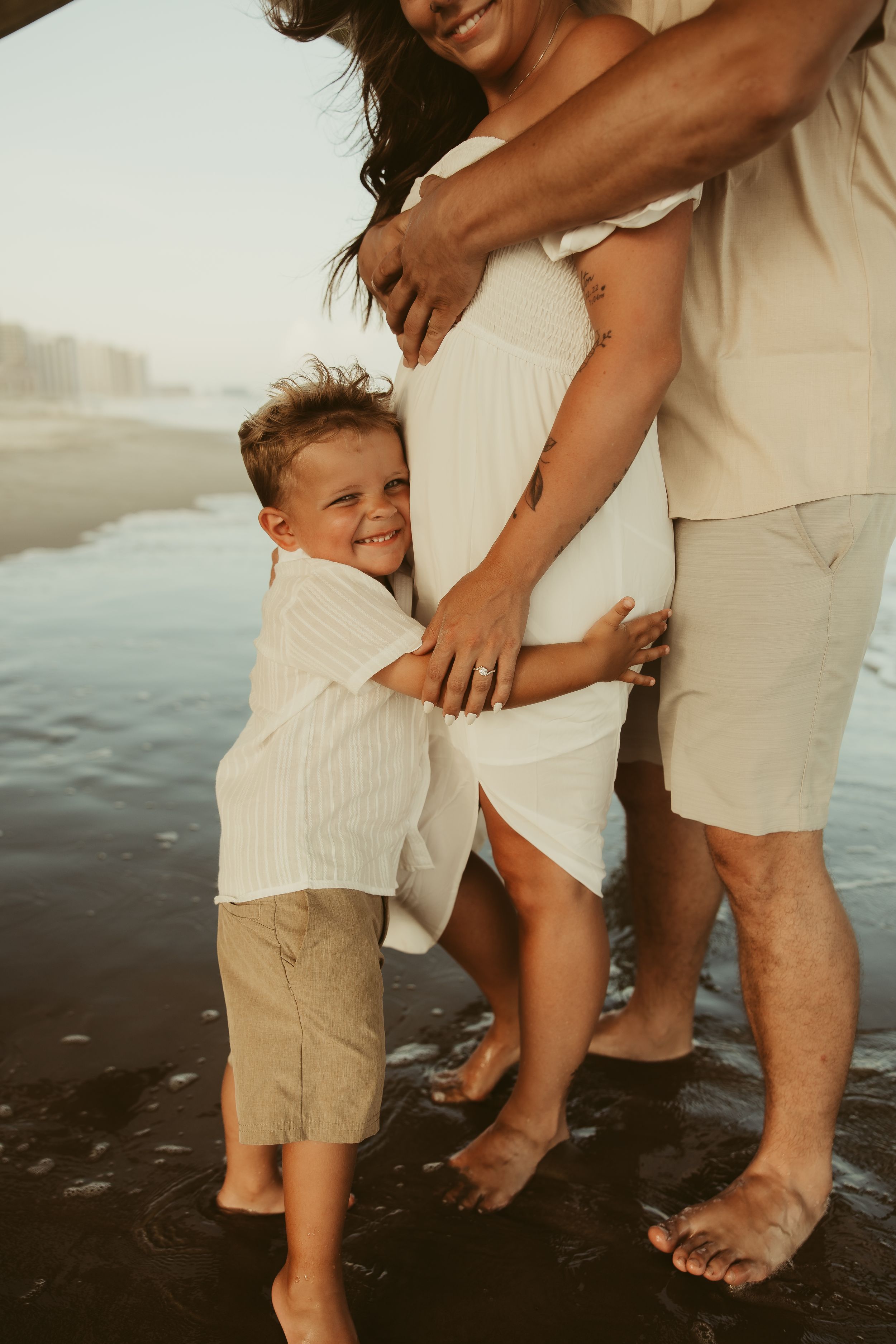 Young boy hugging pregnant mothers belly while family stands together on a beach.
