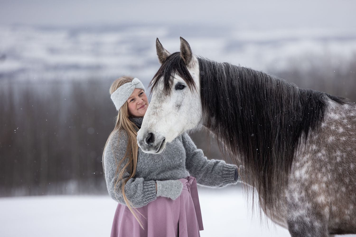Woman in winter clothing shares tender moments with majestic white and black horse in snowy winter landscape.