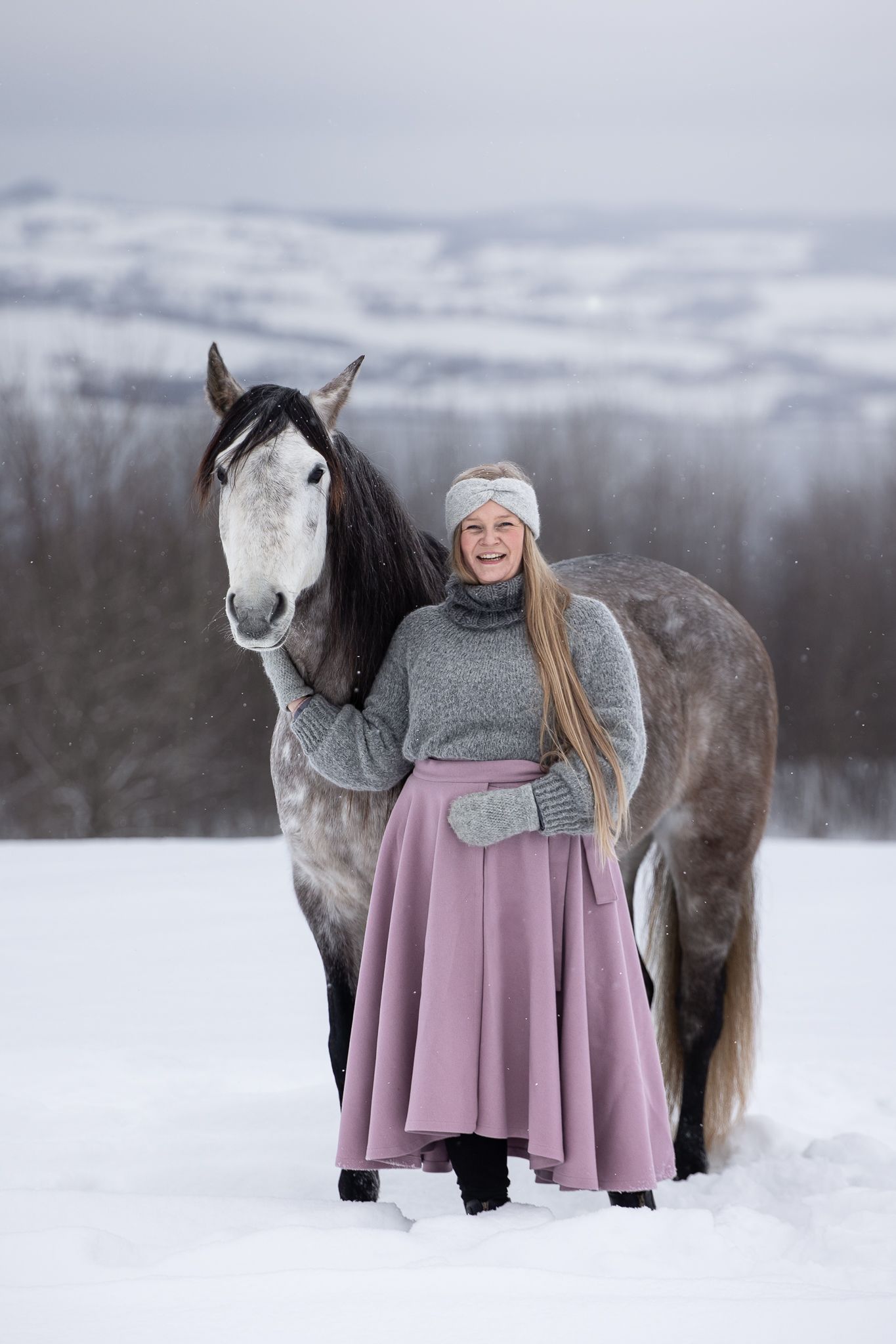 A person in winter attire stands with a gray horse in a snowy landscape wearing a pink poncho and white knit hat.