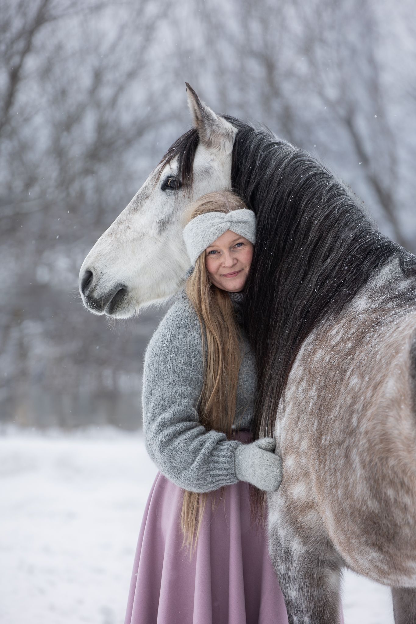 A woman in a pink dress and grey sweater embraces a dappled grey horse in a snowy winter landscape.