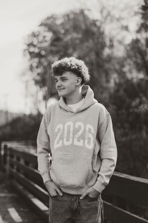 Black and white photo of smiling teen wearing class of 2026 hoodie on wooden bridge.