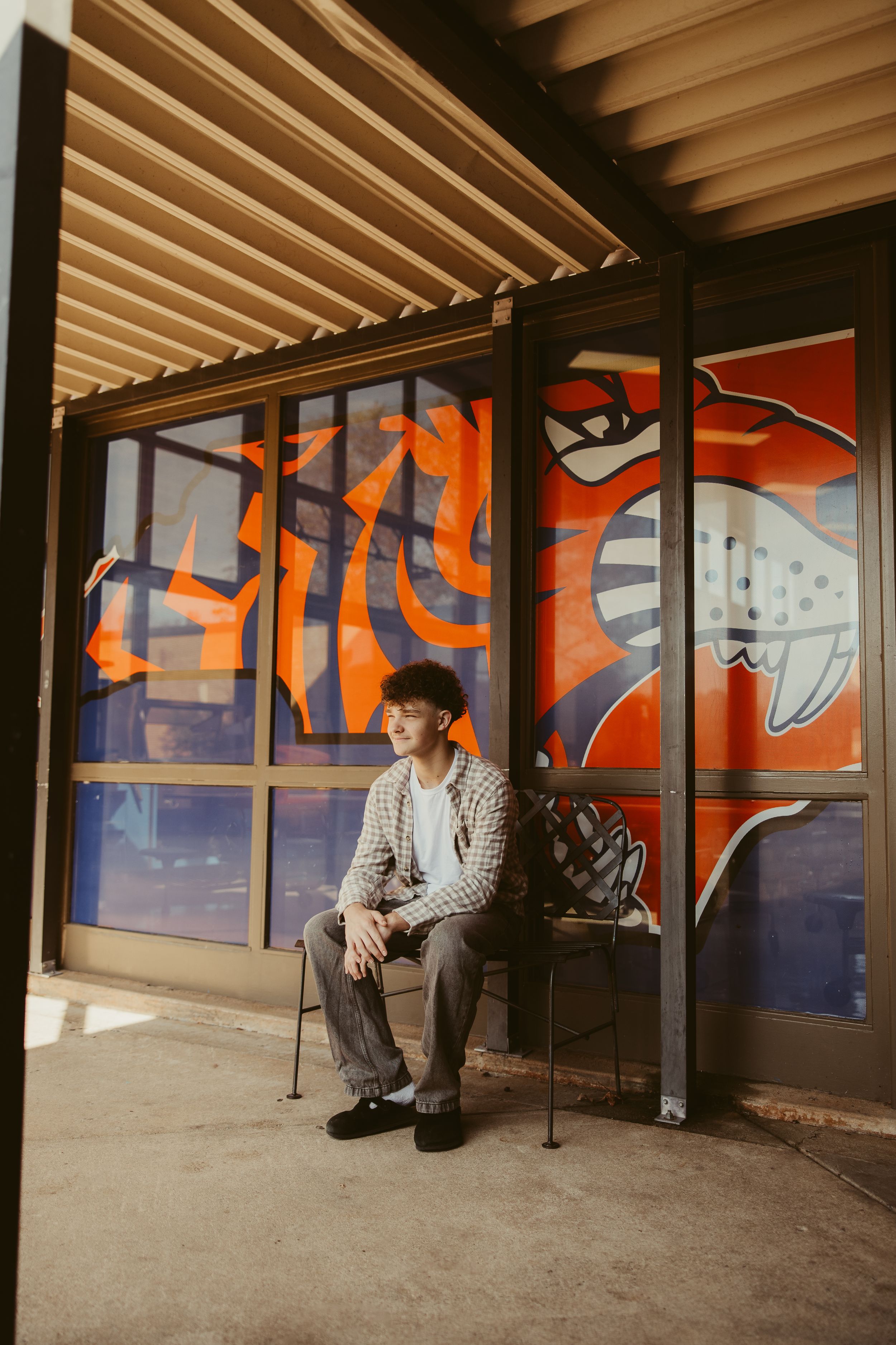 Young man sitting on chair outside building with colorful orange graffiti mural on windows behind him.