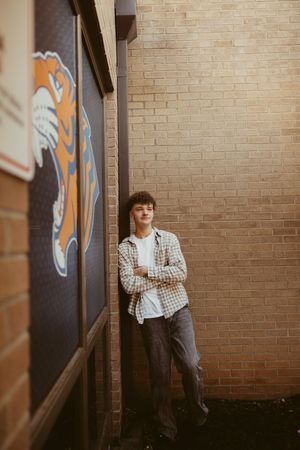 Teen boy in plaid shirt leaning against brick wall next to school mascot tiger mural.
