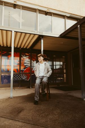 Young man in plaid shirt sitting on stool under modern building overhang with colorful mural.