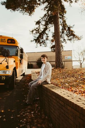 Smiling teen boy sitting on brick wall beside yellow school bus number 79 in autumn.