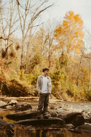 Young man standing on rocks in autumn creek surrounded by colorful fall foliage trees.