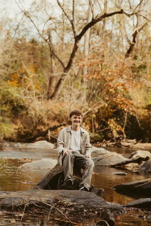 Smiling teen boy sitting on rocks by a peaceful creek in golden autumn forest setting.