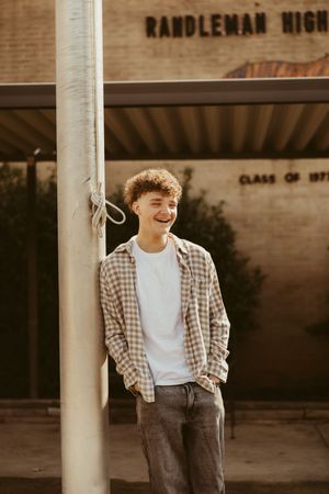 Laughing teenage boy with curly hair leaning on flagpole outside Randleman High School building.