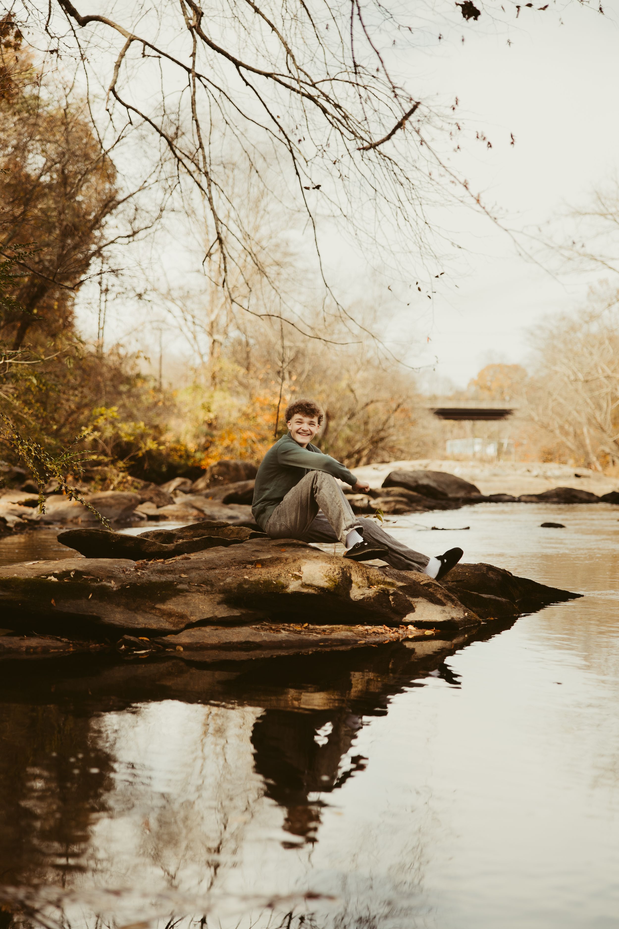 Smiling teen sitting on rocks by calm reflective creek surrounded by bare autumn trees.