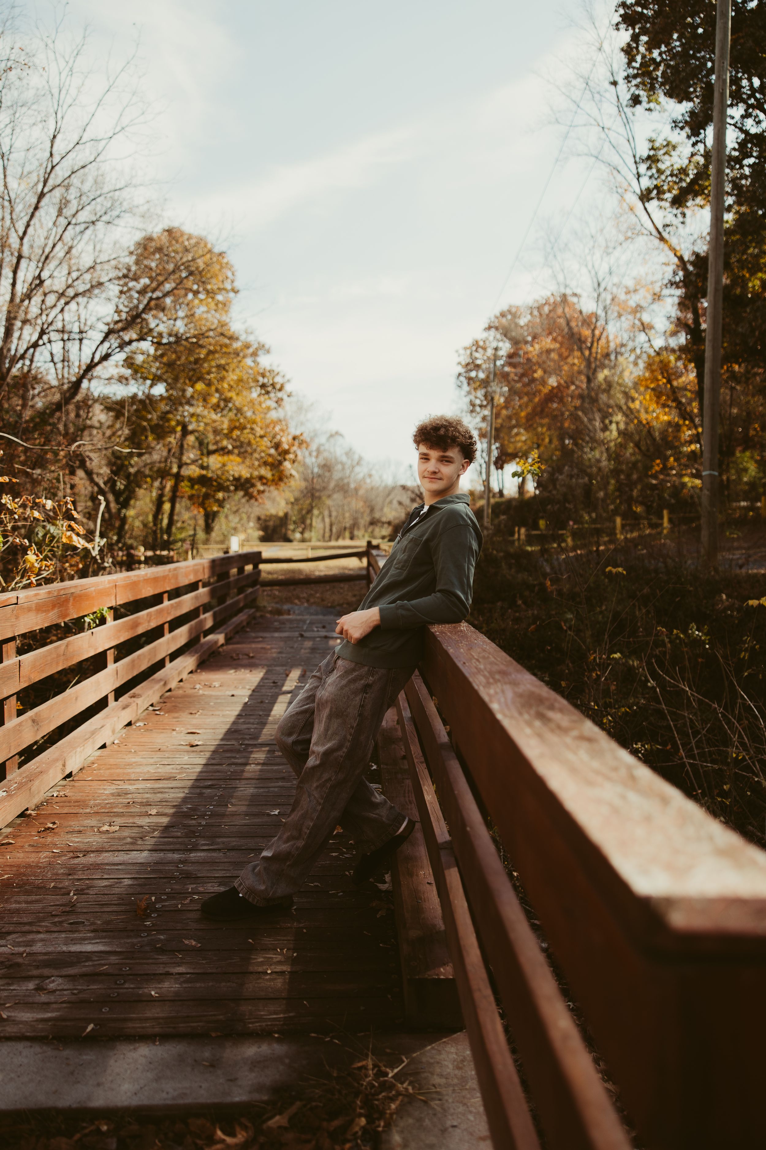 Teen boy leaning on wooden bridge railing in autumn park with colorful fall trees.
