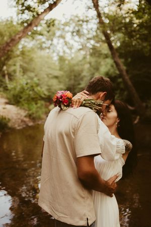 Bride and groom embrace in a forest creek, bride holding a vibrant colorful bouquet in a white dress.