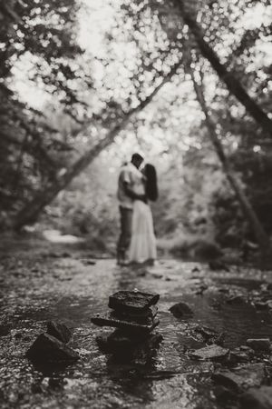Black and white artistic photo of couple embracing in a forest creek with stacked stones in the foreground.