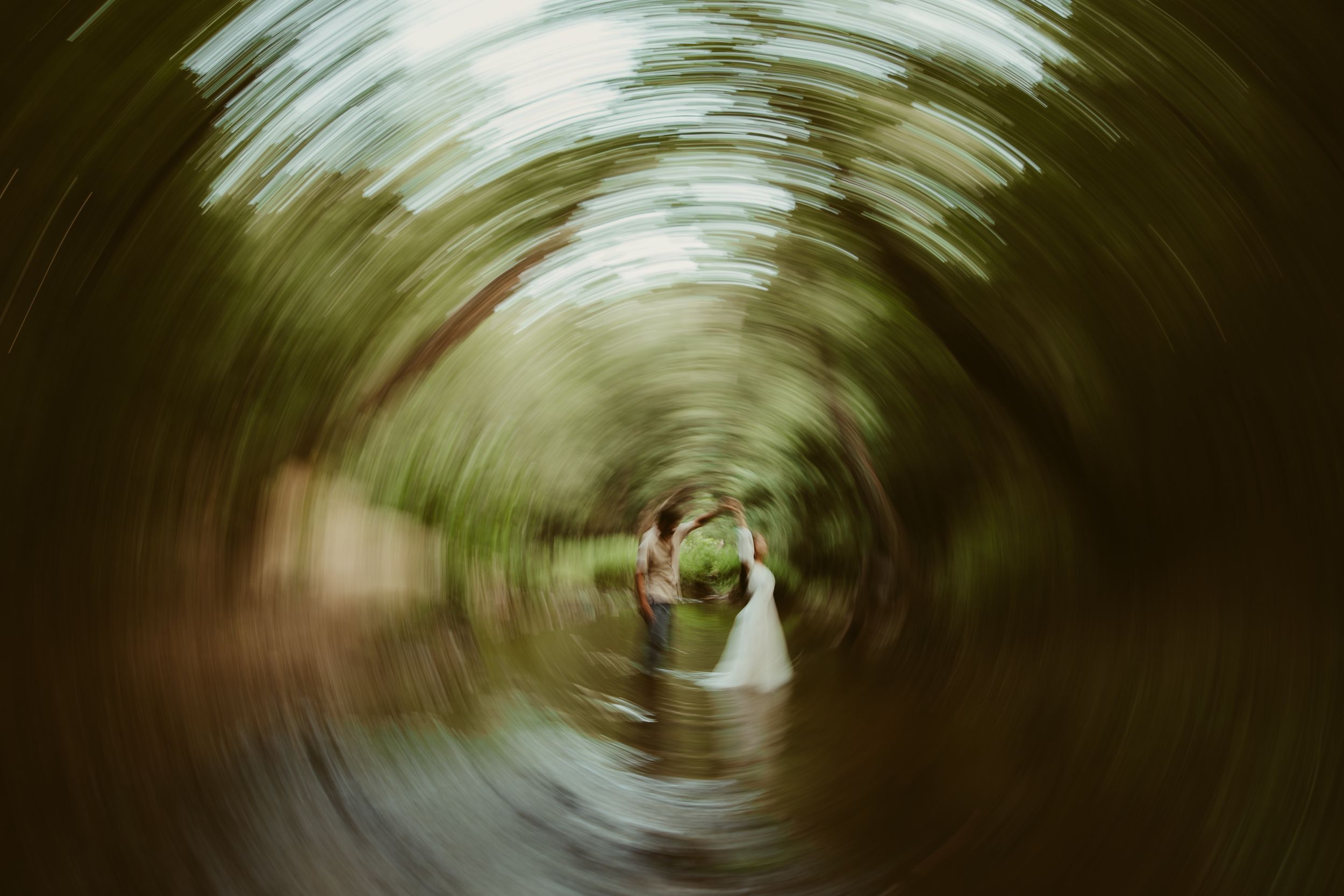 Creative swirl effect photo of bride and groom standing in a lush green forest creek on wedding day.
