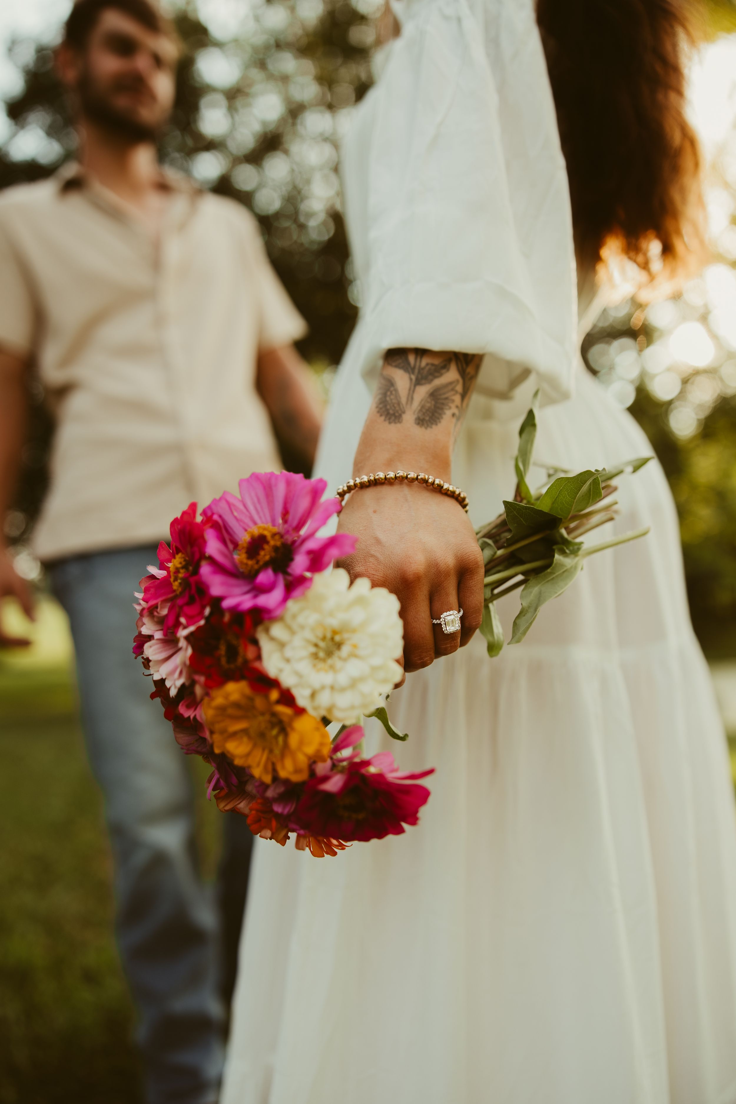 Close-up of bride in white dress holding colorful zinnia bouquet with engagement ring and floral tattoo visible.