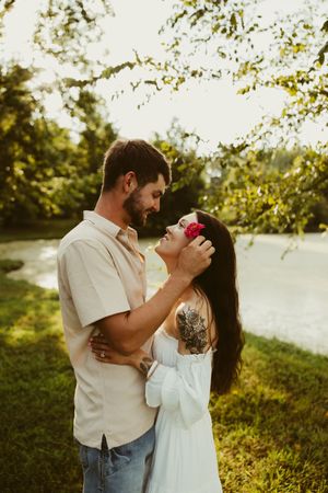 Couple sharing a tender moment under green trees by a river at golden hour, woman with flower in dark hair.