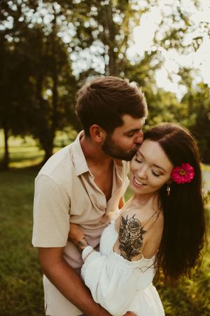 Man kissing woman on temple outdoors at sunset, woman smiling with pink flower in hair and floral chest tattoo.
