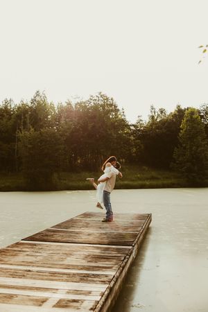 Man lifting woman on a wooden dock over a calm lake surrounded by lush green trees at sunset.