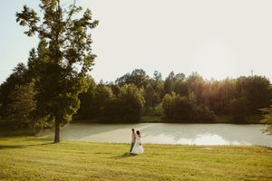 Bride and groom embrace near a sunlit pond surrounded by lush green trees during golden hour outdoor wedding.