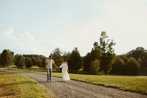 Bride and groom hold hands walking down a gravel path surrounded by green trees on a sunny wedding day.