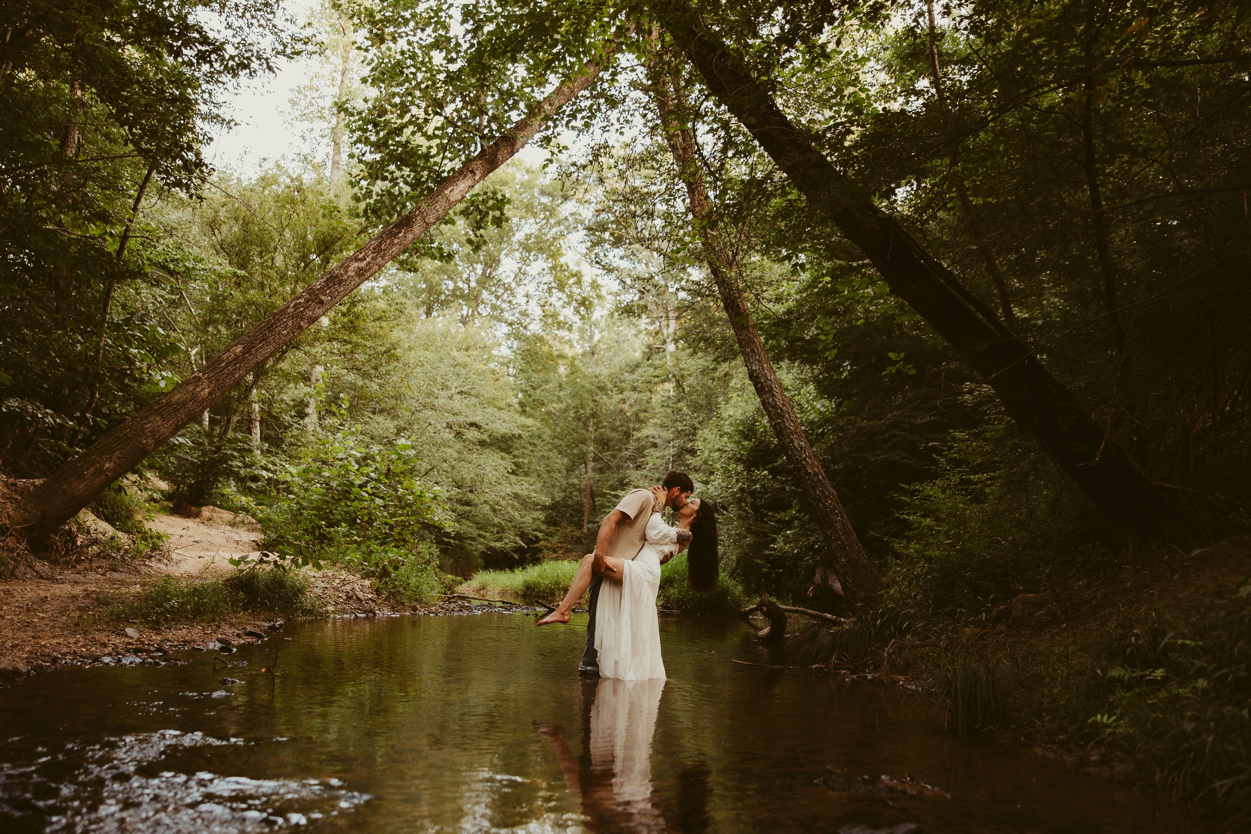 Bride and groom share a romantic embrace while standing in a shallow forest creek surrounded by lush trees.